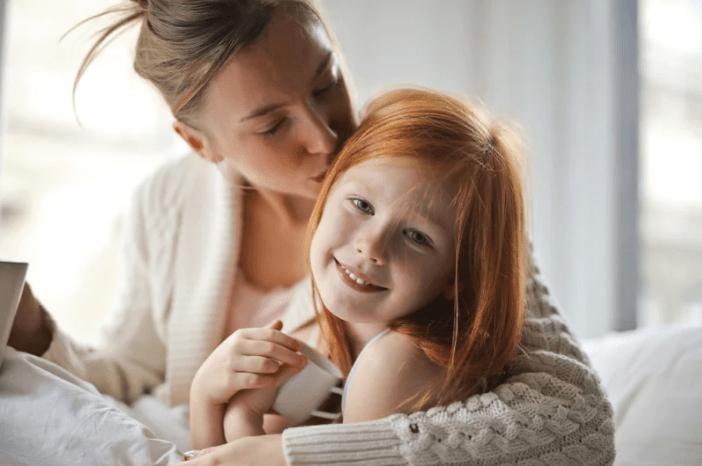 Mother enjoying a mug of coffee while lovingly kissing her daughter on the side of her face. The daughter is smiling, and is holding a mug clasped in both hands to her chest.